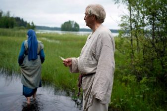 Suomalaisessa perinnemaisemassa nainen ja mies muinaisiin asuihin pukeutuneena.