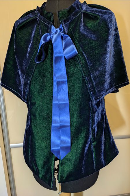 Kuva edestä, linjakas liivi, sinivihreää viskoosisamettia, sininen pitkä rusetti ja hartioilla keeppi.
