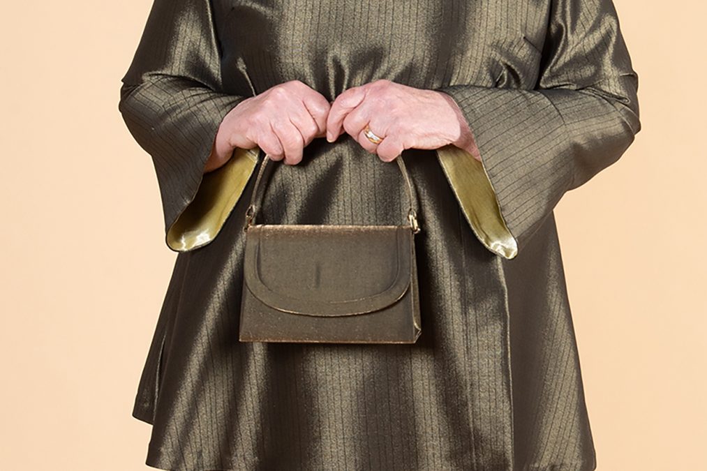 Lähikuvassa kädet pitävät pienen käsilaukun kahvasta, trumpettihihat.