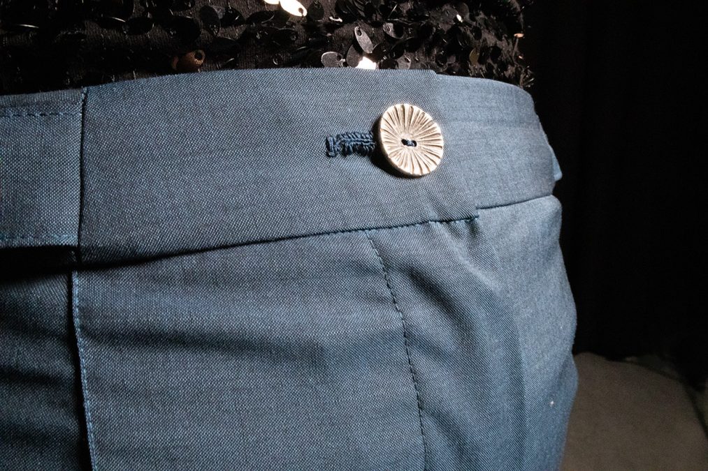 Lähikuva edestä, sinisten villakangashousujen tyylikäs vyötärö ja vaalea nappi.