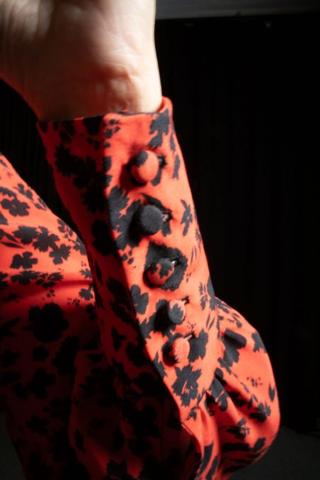 Lähikuvassa puna-mustakuvioisen puseron ranneke, nappirivin napit on päällystetty puserokankaalla.