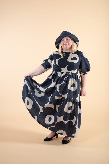 Kuva edestä, käsi avaa leveän frillamekon helmaa sivulle, päässä kesähattu samasta pellavakankaasta.