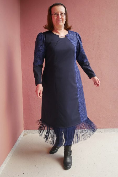 Kuva edestä, sinisen juhlamekon helmassa pitkät hapsut, miehustan vasemmalla puolella kaitale pitsikangasta villakankaan päällä.