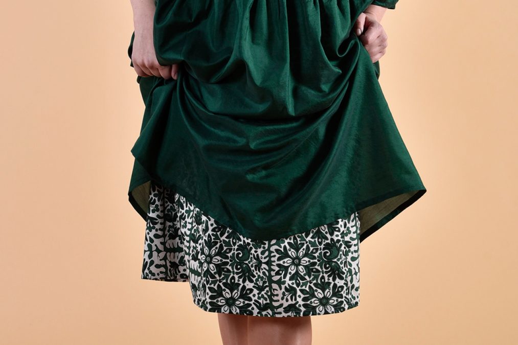 Lähikuvassa helmat, kädet nostavat vihreää päällimekkoa, alta näkyy valkovihreän mekon helma.