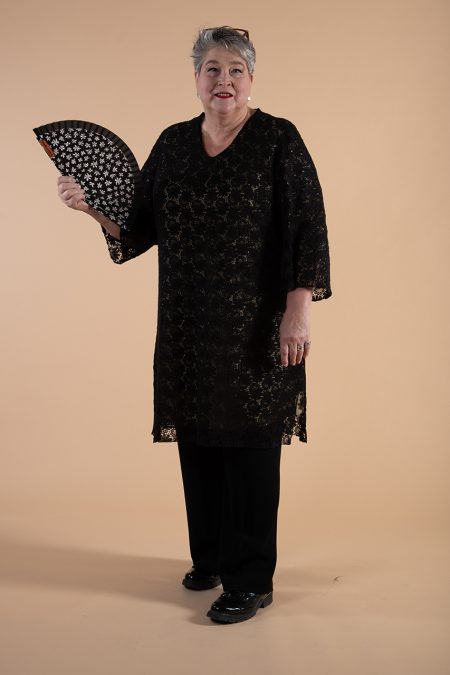 Kuva edestä, mustan pitsitunikan alta väikkyy vaaleaa, kädessä viuhka.